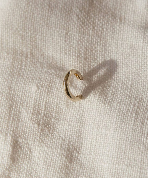 The Huggie, a small 14K gold hoop earring, sits on textured off-white fabric and casts a subtle shadow—ideal for those who love minimalist jewelry.