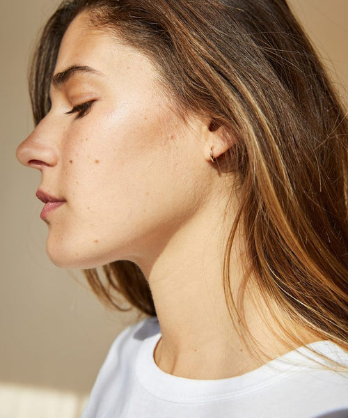 A young woman with long brown hair is shown in profile, eyes closed, wearing a white top and minimalist jewelry—a small 14K gold Huggie earring. Sunlight highlights her natural skin texture and moles.