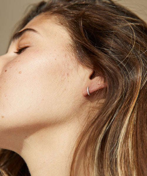 Close-up of a woman in profile with smooth skin, long brown hair, and a delicate Huggie with Diamonds earring. The image highlights her natural makeup and the sleek elegance of these minimalist diamond hoops.