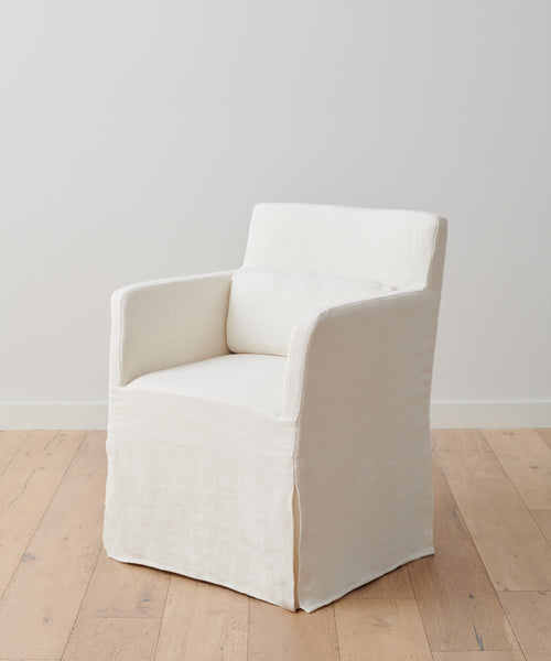 The Sunset Dining Arm Chair Slipcover in alabaster linen, featuring a matching lumbar pillow, rests on a light wooden floor against a plain white wall.