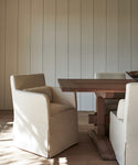 A minimalist dining area features a wooden table and Sunset Dining Arm Chair Slipcovers in alabaster linen, set against a wall with vertical paneling and natural wood flooring.