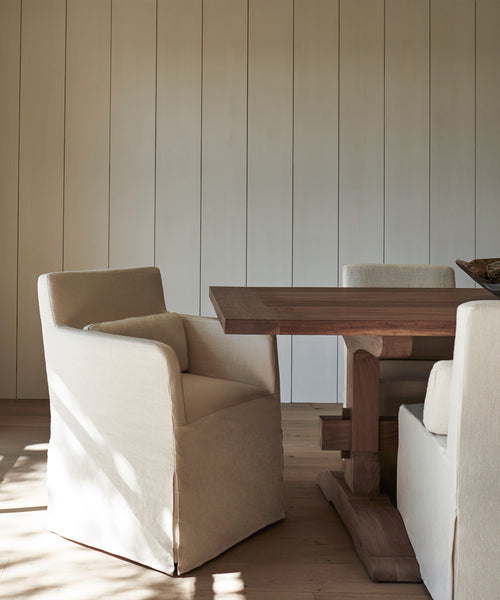 A minimalist dining area features a wooden table and Sunset Dining Arm Chair Slipcovers in alabaster linen, set against a wall with vertical paneling and natural wood flooring.