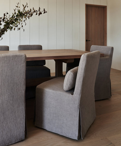 A modern dining room features a wooden table with six chairs in Sunset Dining Chair Slipcovers (ash linen) on a light wood floor. A leafy branch decorates the tabletop, and a wooden door appears in the background.