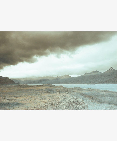Awash 5 Framed Print features a dramatic, rocky landscape with a river under dark clouds and distant mountains. This limited edition artwork is printed on museum-quality cotton rag paper, capturing a moody, desolate atmosphere.