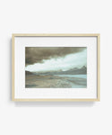 The Awash 5 Framed Print is a limited edition photograph featuring a wide, barren landscape with rocky terrain, distant mountains, and a cloudy sky, presented in a light wood frame with a white mat.