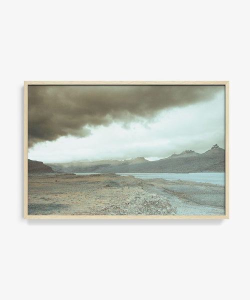 The Awash 5 Framed Print is a limited edition museum-quality cotton rag photograph depicting distant mountains and calm water under a cloudy sky, with muted colors that evoke a moody, atmospheric landscape.