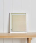 The Untitled 2 Framed Print, a limited edition with a white frame and beige/light tones, is shown on a light wooden shelf against a white, vertically paneled wall.