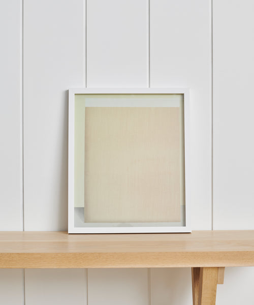 The Untitled 2 Framed Print, a limited edition with a white frame and beige/light tones, is shown on a light wooden shelf against a white, vertically paneled wall.