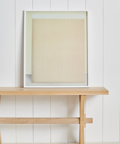 The Untitled 2 Framed Print by Tycjan Knut, featuring soft beige and cream tones, rests on a simple wooden bench against a white paneled wall—an elegant accent inspired by Jenni Kayne Home.