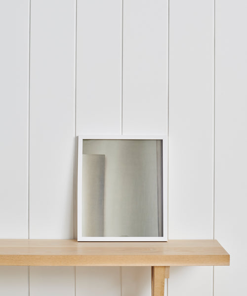 A square mirror with a white frame sits on a light wooden shelf against a white paneled wall, styled alongside the Untitled 5 Framed Print for an elevated Jenni Kayne Home aesthetic.