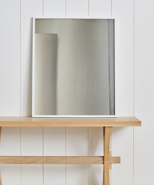 The Untitled 5 Framed Print with a white frame sits on a light wooden table against a white paneled wall, reflecting the refined aesthetic of Jenni Kayne Home. The print reflects a blank wall and part of another textured surface.