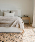 A cozy, minimalist bedroom features a white bed with beige bedding, neutral pillows, a wooden nightstand with a bowl and candle, and the Wilder Diamond Jute Rug on the floor as sunlight streams through the curtains.