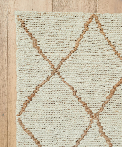 A close-up of the Wilder Diamond Jute Rug in cream, featuring a brown diamond pattern. The image highlights the rug’s corner on a light wooden floor, showcasing its rich texture and intricate handwoven details.