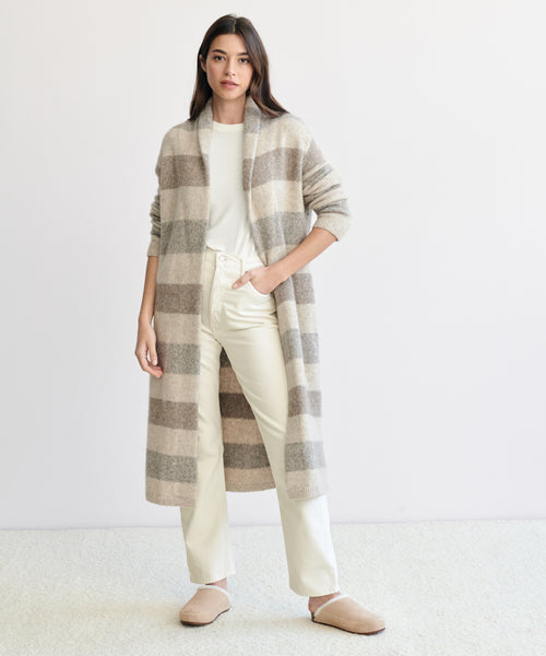 A woman with long dark hair and a neutral expression stands before a light background, wearing a beige and gray striped cardigan, white t-shirt, 90s Pinch Waist Jean, and beige slippers.