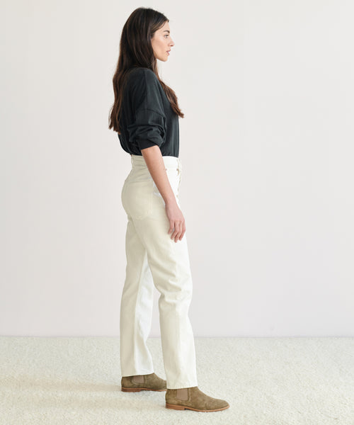 A woman with long dark hair stands in profile against a plain background, wearing a black long-sleeve top, 90s Pinch Waist Jean in white, and brown boots.