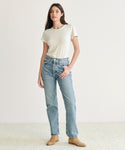 A woman with long dark hair and one hand in her pocket wears a short-sleeved white t-shirt, the 90s Pinch Waist Jean in light blue organic cotton denim, and tan suede ankle boots against a plain light background.