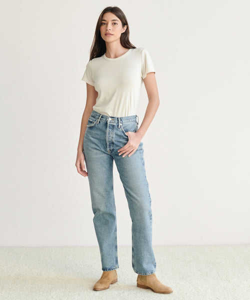 A woman with long dark hair and one hand in her pocket wears a short-sleeved white t-shirt, the 90s Pinch Waist Jean in light blue organic cotton denim, and tan suede ankle boots against a plain light background.
