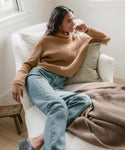 A woman in a tan ribbed sweater and 90s Pinch Waist Jeans relaxes on a white armchair, her head resting on her hand as she smiles. A brown textured blanket and cushion are beside her, with natural light brightening the room.