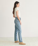 A woman stands in profile against a plain background, wearing a white short-sleeve shirt, light blue organic cotton 90s Pinch Waist Jean, and tan shoes.