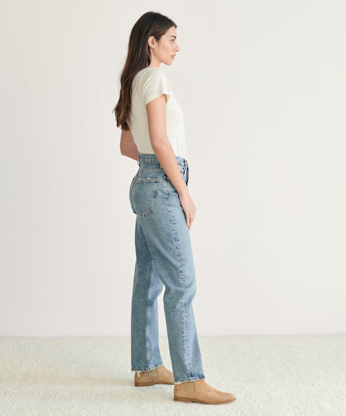 A woman stands in profile against a plain background, wearing a white short-sleeve shirt, light blue organic cotton 90s Pinch Waist Jean, and tan shoes.