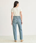 A person with long brown hair, wearing a white short-sleeve tee and 90s Pinch Waist Jean in organic cotton denim, stands barefoot facing away from the camera against a plain white background.