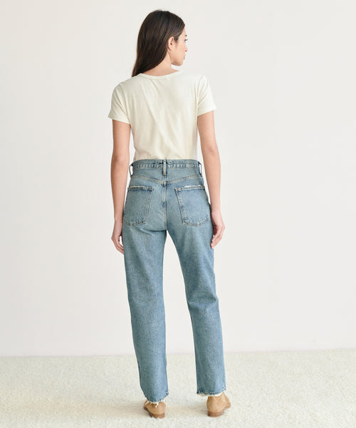 A person with long brown hair, wearing a white short-sleeve tee and 90s Pinch Waist Jean in organic cotton denim, stands barefoot facing away from the camera against a plain white background.