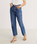 Model wears a white t-shirt tucked into high-waisted 90s Crop Jeans in vintage blue with hands in pockets, paired with tan strappy sandals, standing on a light carpet against a plain background.