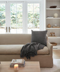 A cozy living room with a beige sofa is accented by a dark gray Alpaca Basketweave Pillow and matching throw, facing large windows. A lit candle and books rest on a wooden coffee table, with shelves in the background.