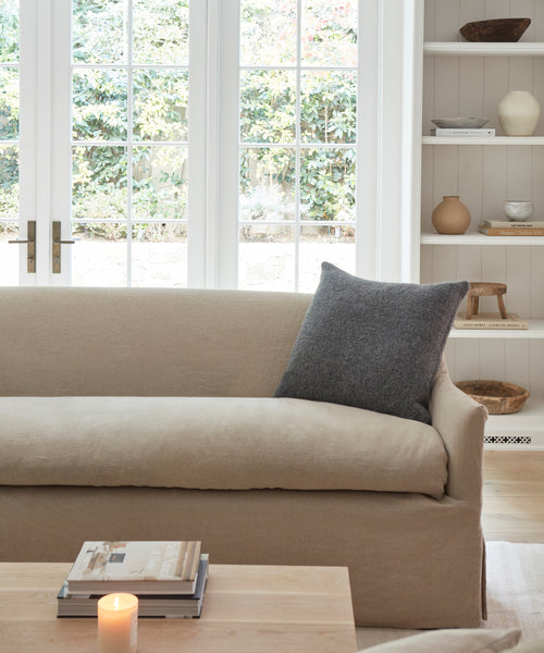 A cozy living room with a beige sofa, an Alpaca Basketweave Pillow in dark gray, a lit candle on a wooden coffee table with books, built-in shelves with decor, and large glass doors revealing greenery outside.
