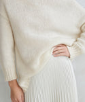 A person wearing the Alpaca Cocoon Crewneck, a loose cream-colored chunky sweater, paired with a white pleated skirt. The cropped photo shows only the torso and upper legs, with one hand resting on their hip.
