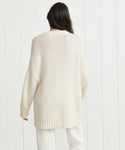A person with long dark hair stands facing away, wearing the Alpaca Cocoon Crewneck in cream and white pants against a white paneled wall.