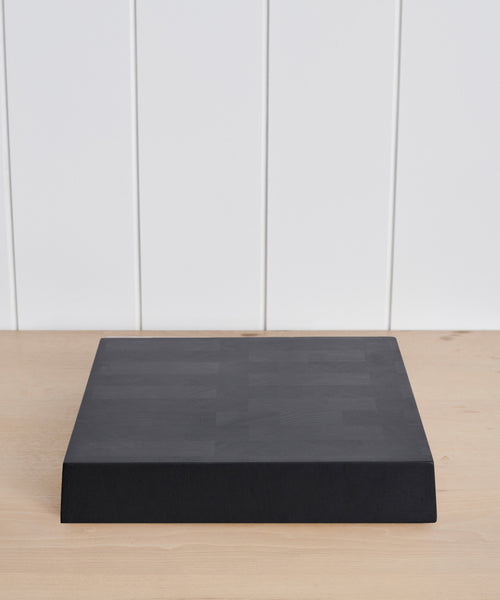 The Square Butcher Block, a large black wooden cutting board, sits on a light wood surface with a white paneled wall in the background.