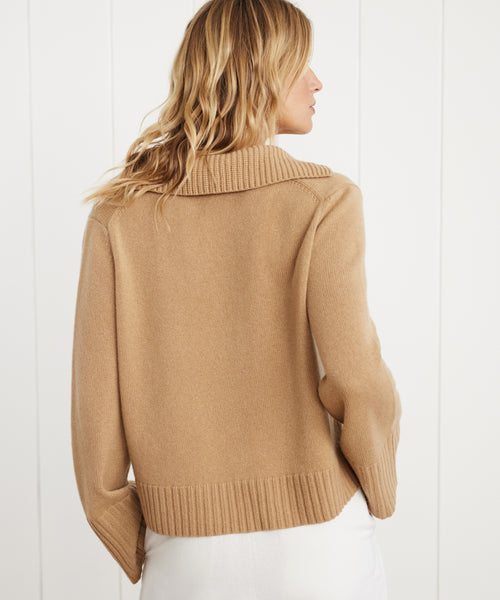 A woman with wavy blonde hair models the Cashmere Frances Polo, a relaxed-fit camel sweater with a wide collar, standing with her back to the camera against a white paneled backdrop.