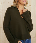 A woman wears the Cashmere Frances Polo, a dark olive relaxed fit knit sweater with a V-neckline, paired with blue jeans as she stands before a light wooden wall. Her face is partially out of frame.