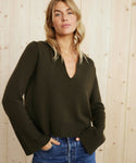 A woman with wavy blonde hair stands in front of a light wooden wall, hands in her pockets, wearing the Cashmere Frances Polo in dark olive with blue jeans. She has a relaxed expression and the sweater features wide ribbed cuffs.