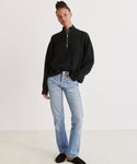 A person stands against a plain background wearing the Cashmere Half Zip sweater in black with a relaxed fit, light blue straight-leg jeans, and black slip-on shoes, one hand in their pocket.