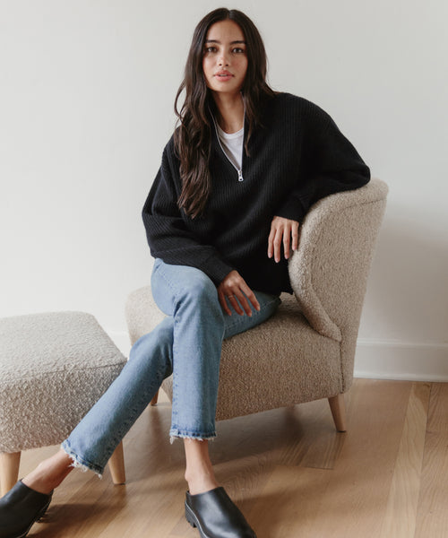 A woman with long dark hair sits on a beige textured chair, wearing the Cashmere Half Zip sweater over a white top, light blue jeans, and black slip-on shoes. She poses relaxedly with one leg crossed and a neutral expression.