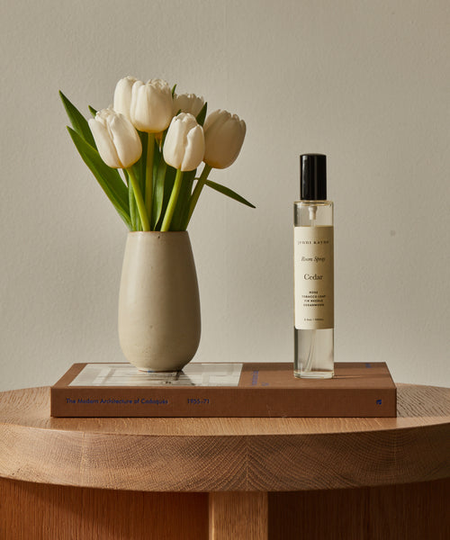 A beige vase with white tulips and a bottle of Cedar Room Spray sit atop a closed architecture book on a wooden surface.
