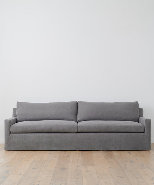 The Harbor Sofa, handcrafted in Los Angeles, features a minimalist charcoal linen design with two large seat and back cushions, set against a white wall on light wooden flooring for a refined, modern look.
