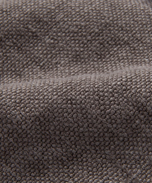 Close-up of the Cove Bed Slipcover shows a textured gray fabric with a slightly raised, bumpy surface and tightly woven fibers. The material appears soft and flexible, forming gentle folds that create subtle shadows.