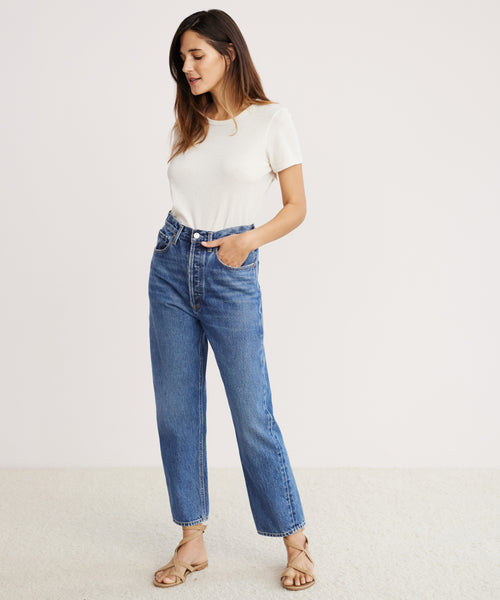 A woman stands against a plain light background, wearing a white short-sleeve t-shirt, strappy sandals, and the 90s Crop Jean. One hand is in her pocket as she smiles softly and gazes slightly downward.