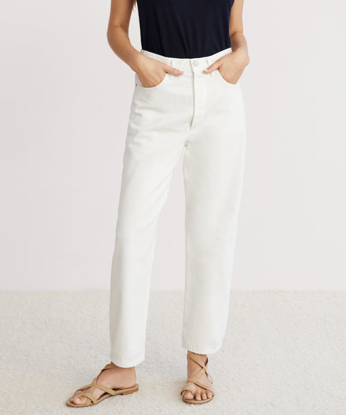 A person stands with hands in the pockets of 90s Crop Jeans, wearing a sleeveless black top and tan sandals, against a plain light background.