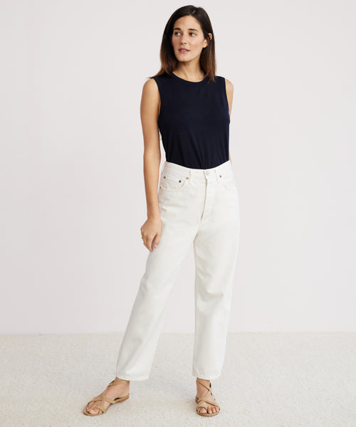 A woman with straight, shoulder-length brown hair stands against a light background, wearing the 90s Crop Jean in white organic cotton, a sleeveless navy top, and beige sandals. She gazes slightly to her left.