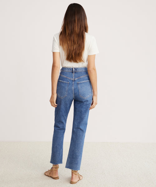A woman with long brown hair wears a white T-shirt, Riley Crop Jean, and sandals. She stands indoors on a light carpet, facing away from the camera with her arms relaxed at her sides.