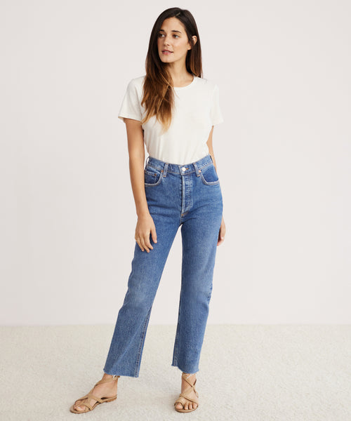 A woman with long brown hair stands against a plain background, wearing a white short-sleeve t-shirt, beige sandals, and high rise Riley Crop Jean made from stretch denim. She looks slightly to the side with a relaxed posture.