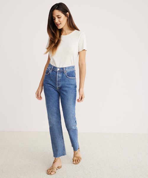 A woman in a white t-shirt and the Riley Crop Jean stands on a light floor against a pale background, wearing beige sandals and looking down with a relaxed expression.