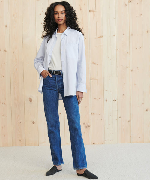 A woman with curly hair stands in front of a light wooden wall, wearing the Boyfriend Shirt in French blue over a white top, paired with blue jeans, a black belt, and black slip-on shoes.