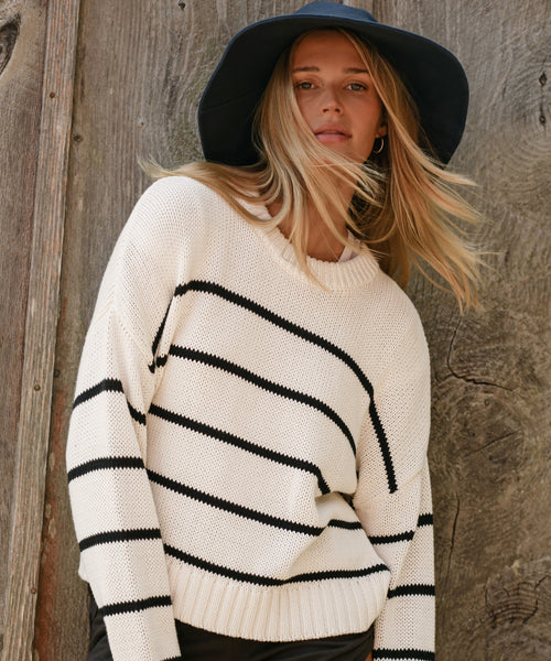 A woman in the Chloe Crewneck, a cropped white knit sweater with black stripes, pairs it with a wide-brimmed navy hat as she stands before a rustic wooden wall, her blonde hair blowing in the wind.