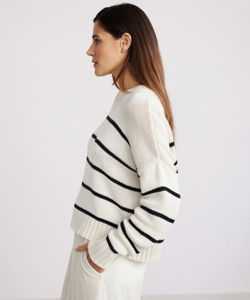 A woman stands in profile against a plain background, wearing the Chloe Crewneck black stripe sweater made from soft Peruvian cotton and cream pants, her hands tucked in her pockets.