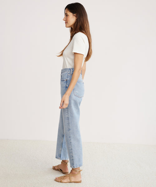Woman in profile against a plain background, wearing a white short-sleeve top, 90s Crop Jean in light blue organic cotton denim by AGOLDE, and tan sandals.
